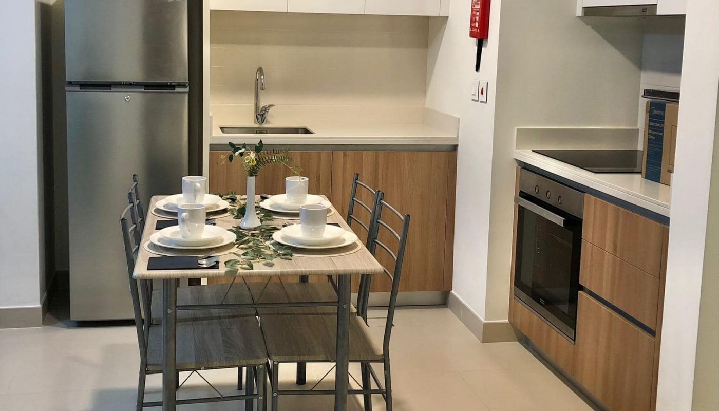 A modern kitchen and dining area in a 1-bedroom flat for sale at Marassi Boulevard, featuring a table set for four, with a refrigerator, sink, oven, and microwave. The space boasts wooden cabinets and neutral-colored countertops and floors.