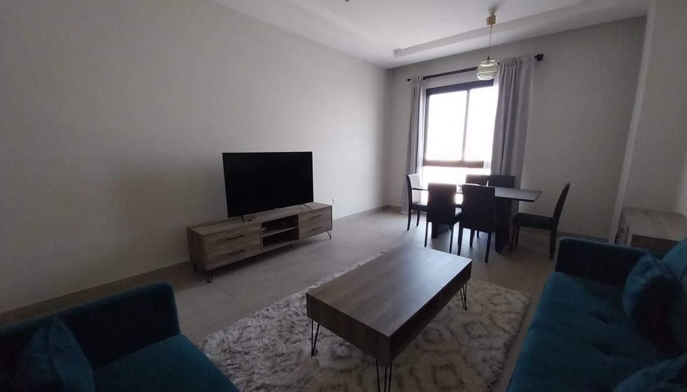 A flat for rent in Hidd features a cozy living room with two blue couches, a wooden coffee table on a rug, and a flat-screen TV on a stand. The space also includes a dining table with four chairs set in front of window curtains.