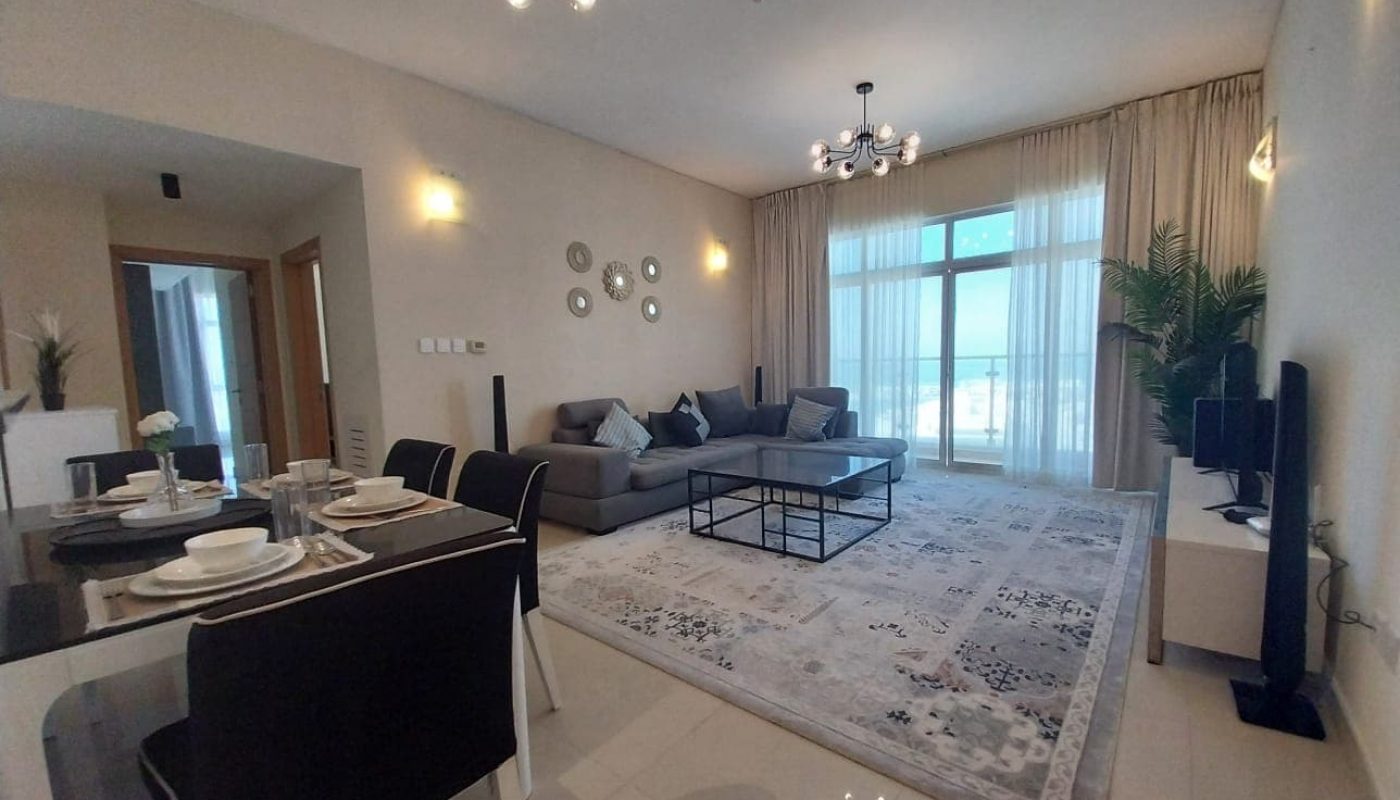 Modern living room with a gray sofa, glass coffee table, wall-mounted TV, dining table set for four, decorative wall plates, large window, and indoor plant.