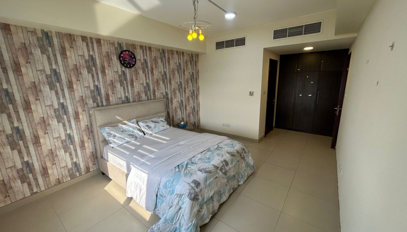 A bedroom with a double bed, patterned duvet, wooden accent wall with a clock, tiled floor, and dark wooden wardrobe near the entrance.