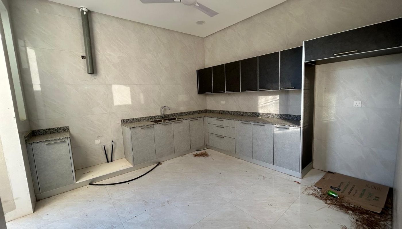 Modern kitchen under construction with unfinished plumbing, loose wires, some debris on the tile floor, and black cabinets mounted on a gray tiled wall.