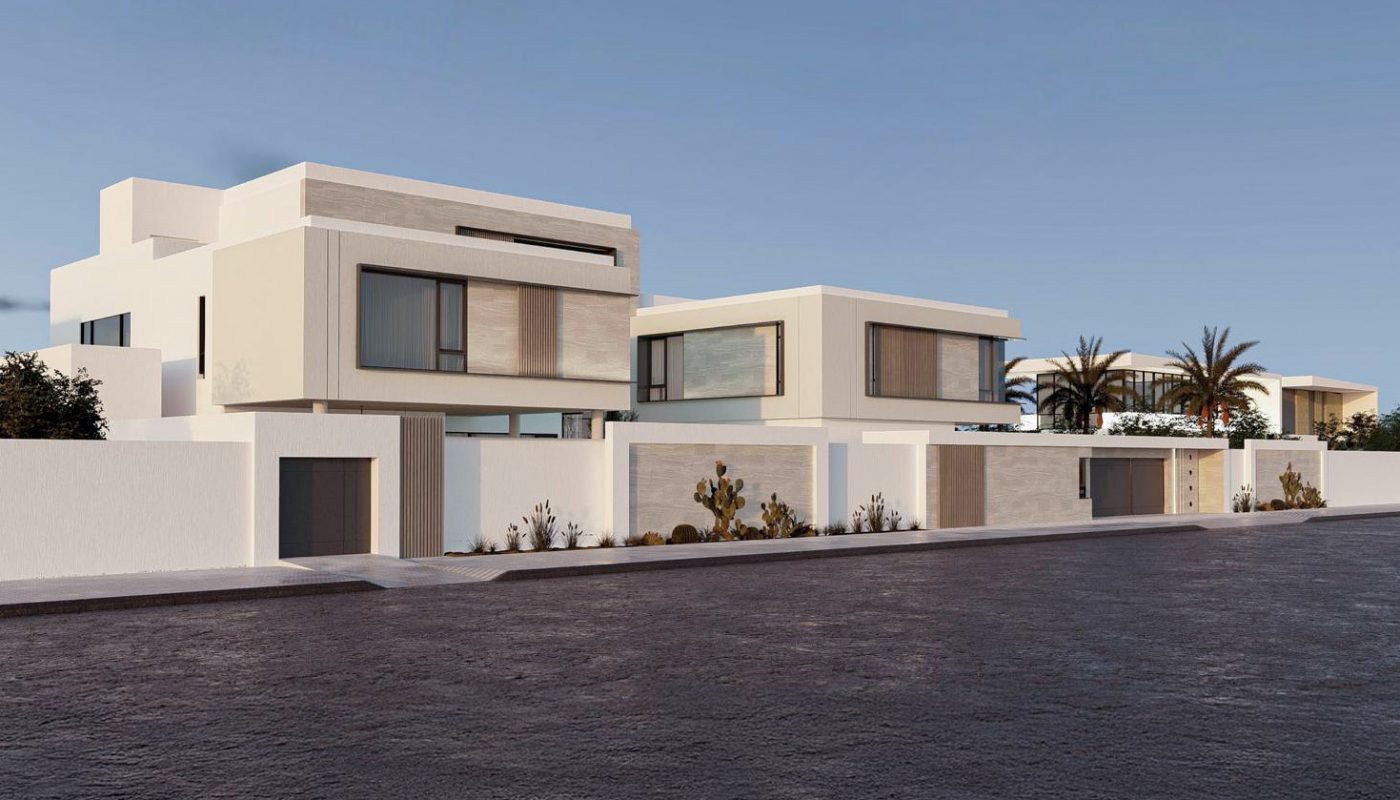Modern white two-story houses with large windows and flat roofs sit along a quiet street bordered by a white wall, with palm trees visible in the background under a clear sky.