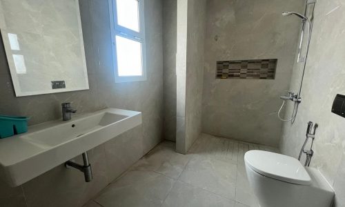 Modern bathroom interior in a flat for rent, featuring a large sink, wall-mounted toilet, and gray tiled walls and floor. Two small windows provide natural light.