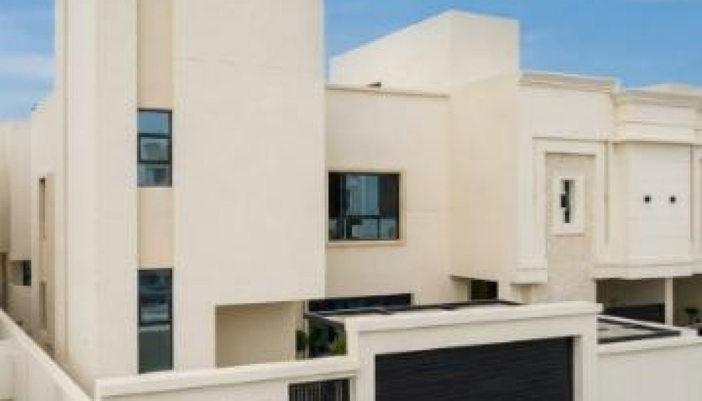 Modern two-story beige house with flat roof, enclosed by a matching perimeter wall, featuring large windows and a double black garage door.