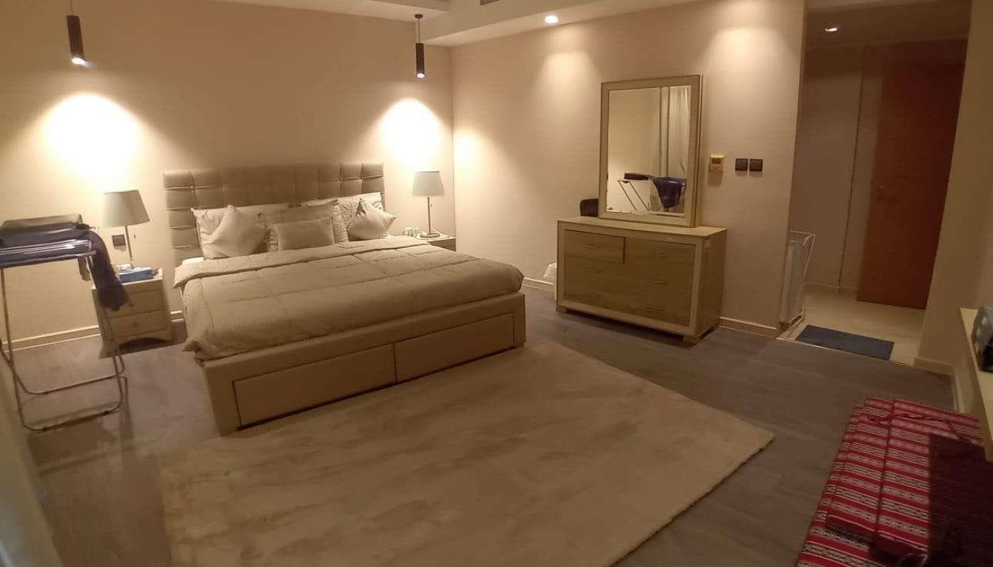 A modern bedroom with a large bed, bedside tables and lamps, a dresser with a mirror, a rug, and folded bedding on the floor. Neutral-colored walls and floor.