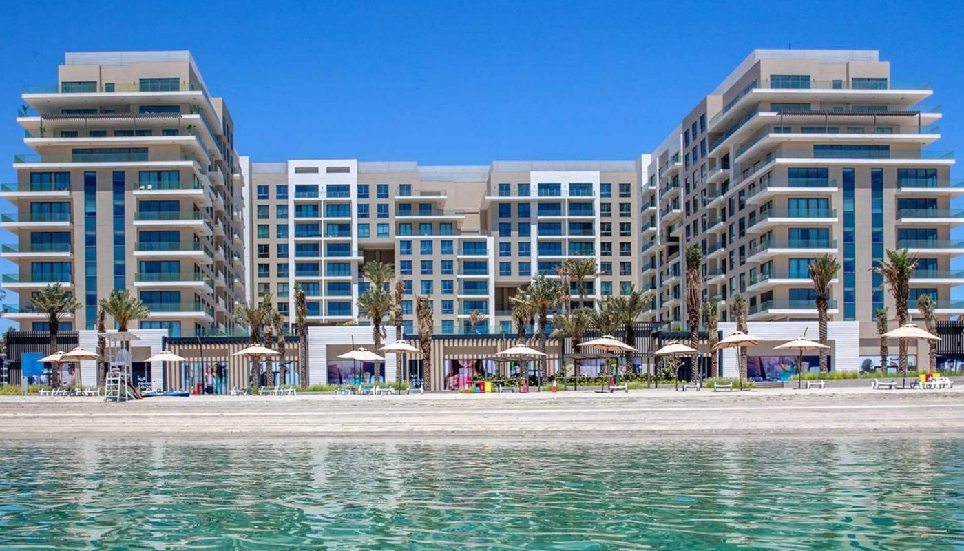 Modern beachfront resort with multiple levels, balconies, palm trees, and sun loungers facing a calm, clear body of water under a clear blue sky.