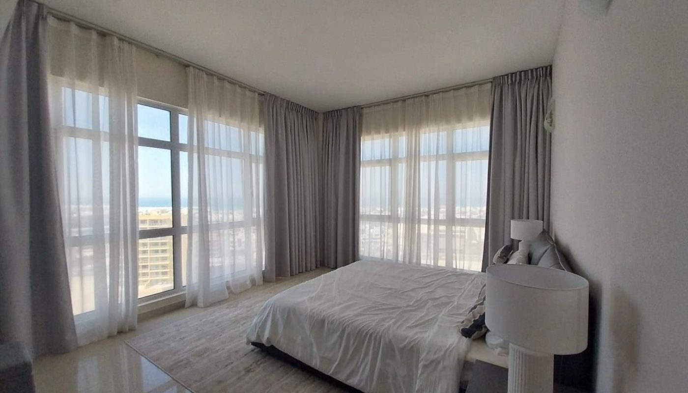 A modern bedroom with large floor-to-ceiling windows, white curtains, a bed with white bedding, a nightstand, and a white lamp.