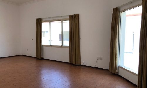 An empty room with tiled flooring, white walls, and mustard-colored curtains on two windows, one on the left and one on the right wall. The room is well-lit with natural light.