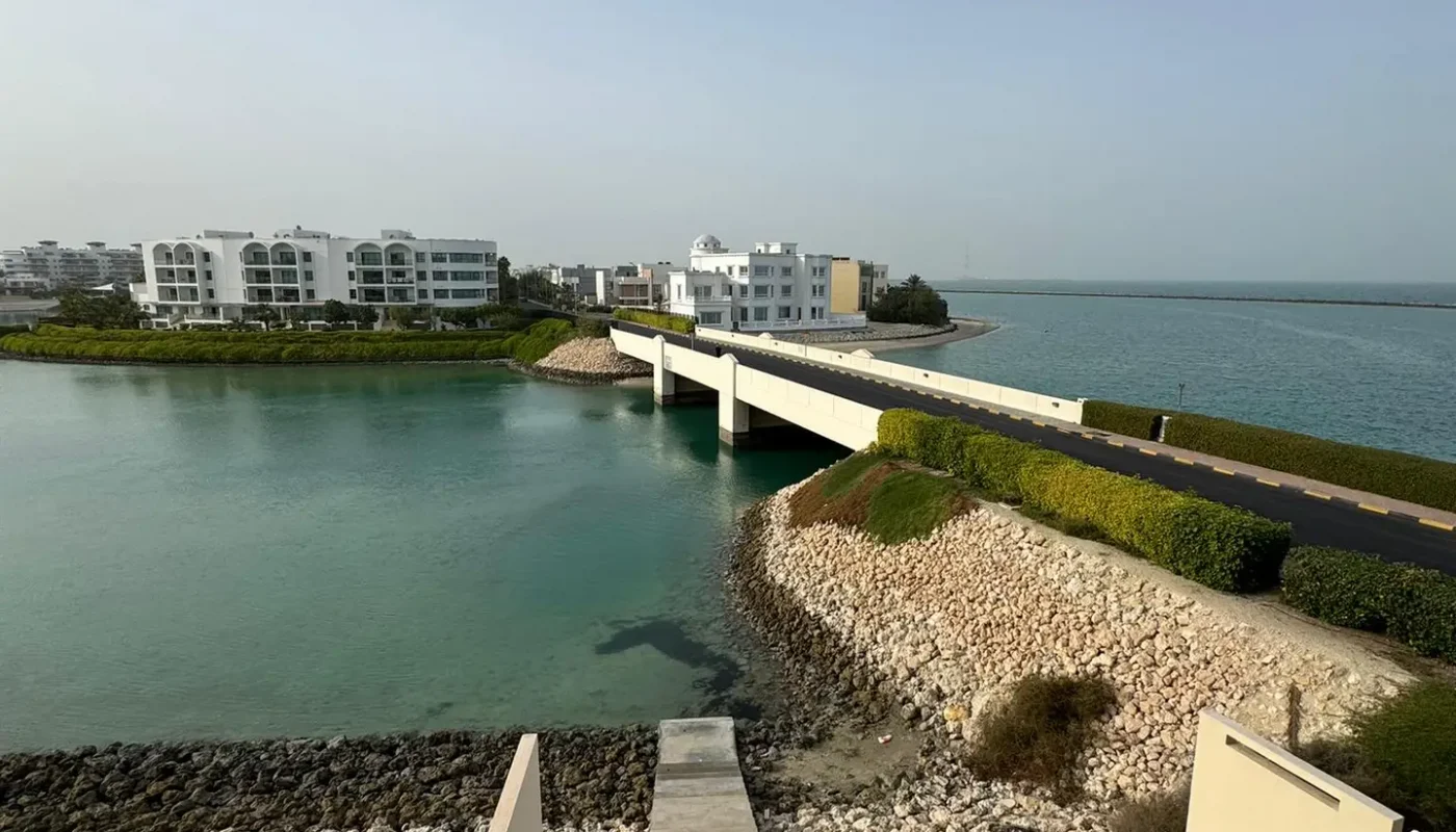 A view of a bridge over turquoise water connects modern white buildings, with a road and rocky shoreline under a clear, blue sky.