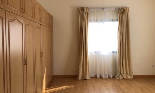 A room with light-colored wooden flooring, a large wardrobe with multiple doors on the left, and a window with sheer white and beige curtains on the right wall.