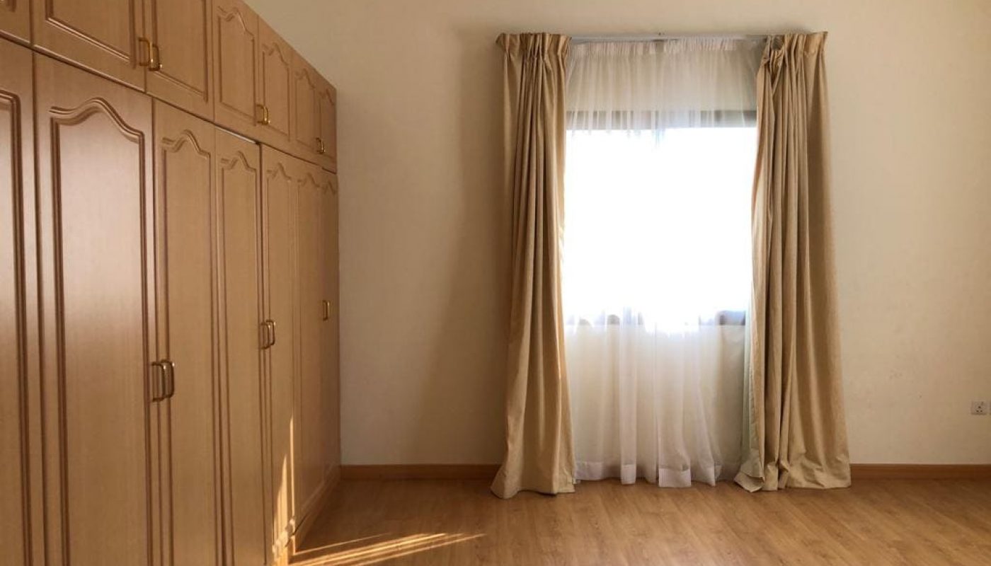 A room with light-colored wooden flooring, a large wardrobe with multiple doors on the left, and a window with sheer white and beige curtains on the right wall.