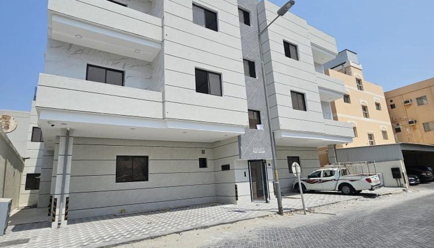 Three-story modern apartment building with balconies and a covered parking area, situated on a paved street under a clear blue sky.