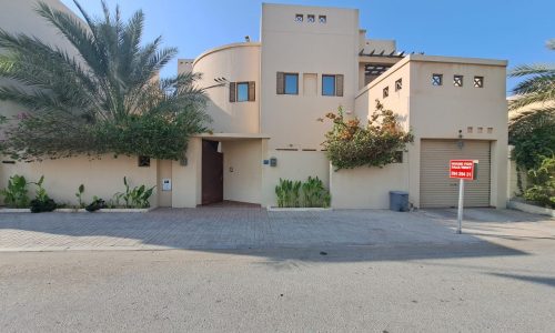 A modern beige villa with a curved design, surrounded by greenery. A "Villa for Sale" sign is posted near the driveway, inviting prospective homeowners to explore this elegant property.