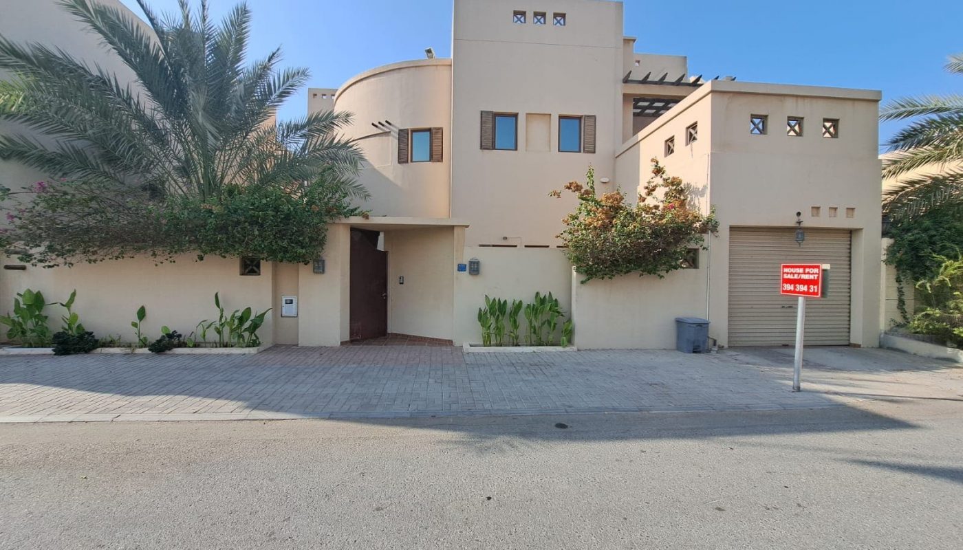 A modern beige villa with a curved design, surrounded by greenery. A "Villa for Sale" sign is posted near the driveway, inviting prospective homeowners to explore this elegant property.