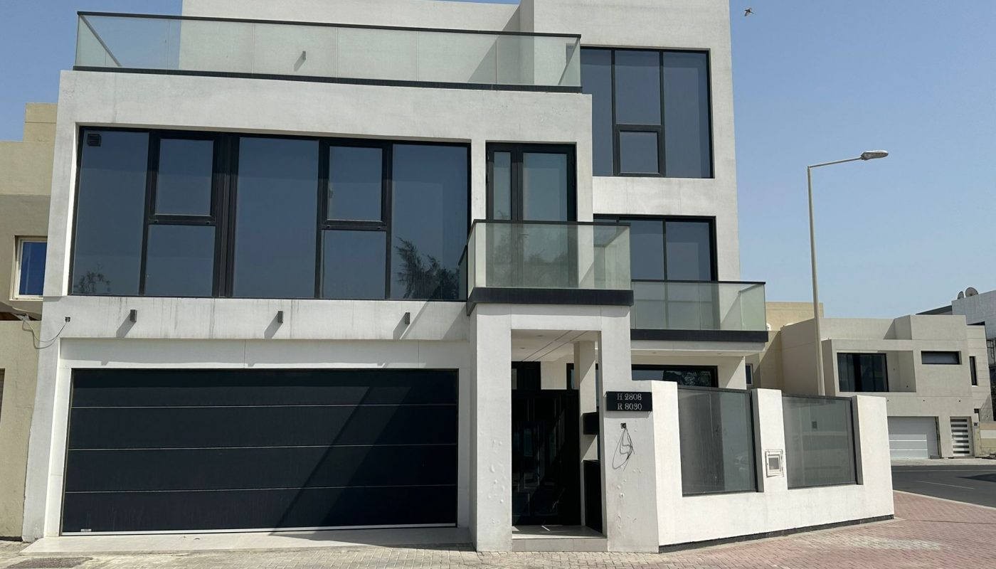 A modern, three-story white house with large black-framed windows, glass balconies, and two garage doors, situated on a paved street under a clear sky.
