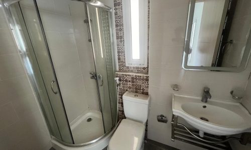 A modern bathroom in a flat for rent, featuring a corner shower stall, toilet, sink, and mirrored cabinet, adorned with white and patterned wall tiles.