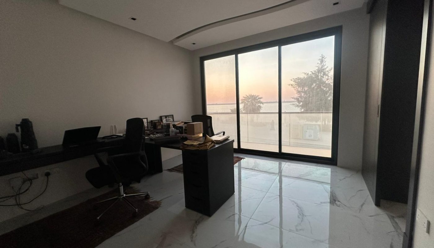 Modern office with two desks and chairs, a central filing cabinet, and large windows showing a view of trees and sunset outside. The room has marble floors and minimalist decor.