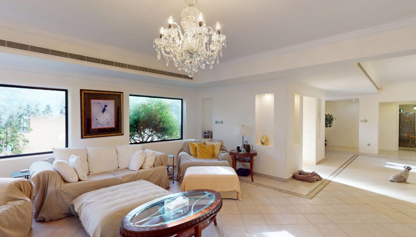 A spacious living room with a chandelier, white sofas, a glass coffee table, large windows, and neutral-toned decor. Natural light fills the room.