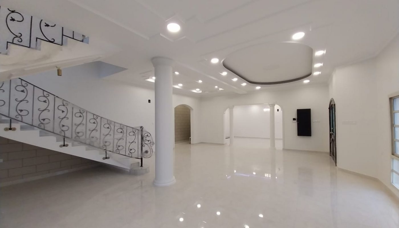Spacious, empty, modern interior with white walls, polished tile floors, decorative ceiling, and a staircase with ornate railing. Multiple recessed lights illuminate the room.