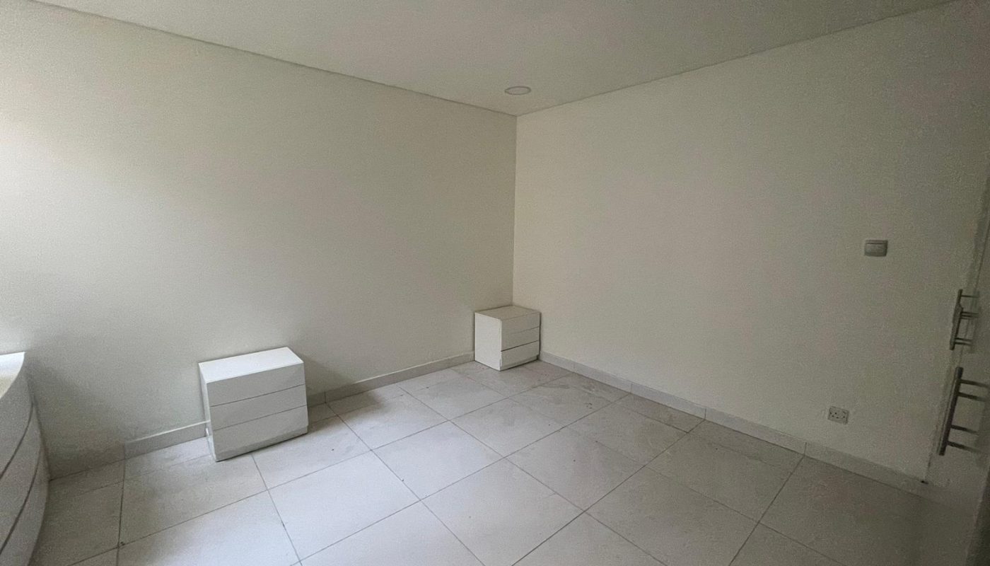 A small, unfurnished room with white tiled floor, off-white walls, two white nightstands, and a dresser near a window. Ceiling has recessed lights.