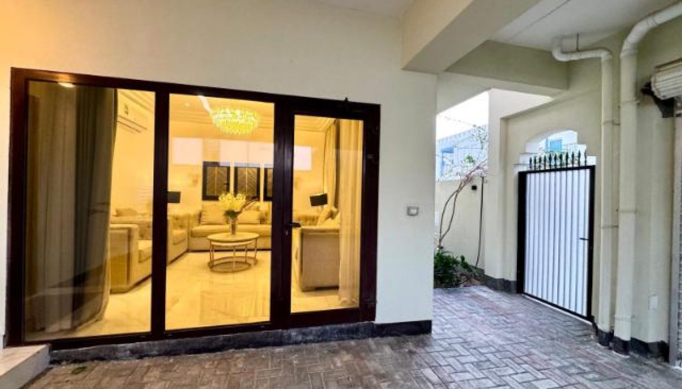 View of a modern living room through large glass doors, with an outdoor paved area and a white gate to the side.