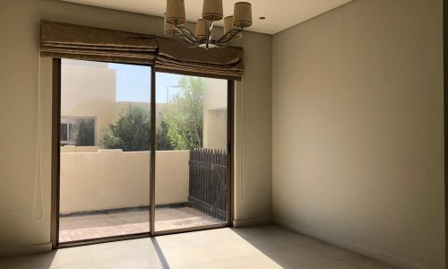 A sunlit empty room with a beige floor and walls, a window with brown curtains, and a modern chandelier. The window opens to a small outdoor area with a wooden fence and greenery visible outside.