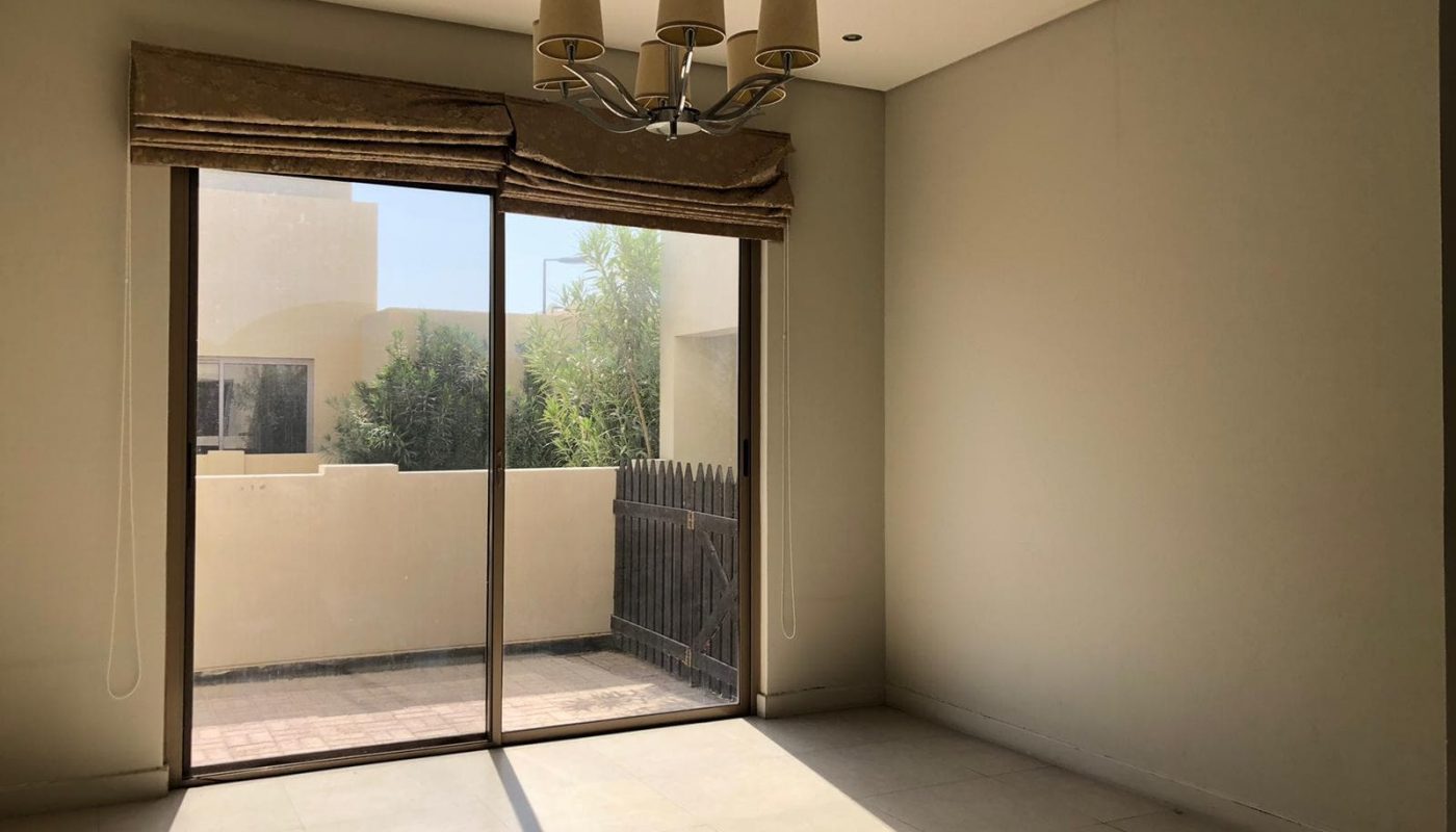 A sunlit empty room with a beige floor and walls, a window with brown curtains, and a modern chandelier. The window opens to a small outdoor area with a wooden fence and greenery visible outside.