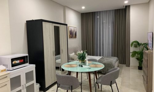 Modern studio apartment interior featuring a dining table with chairs, a bed, and kitchen appliances against neutral-toned decor.