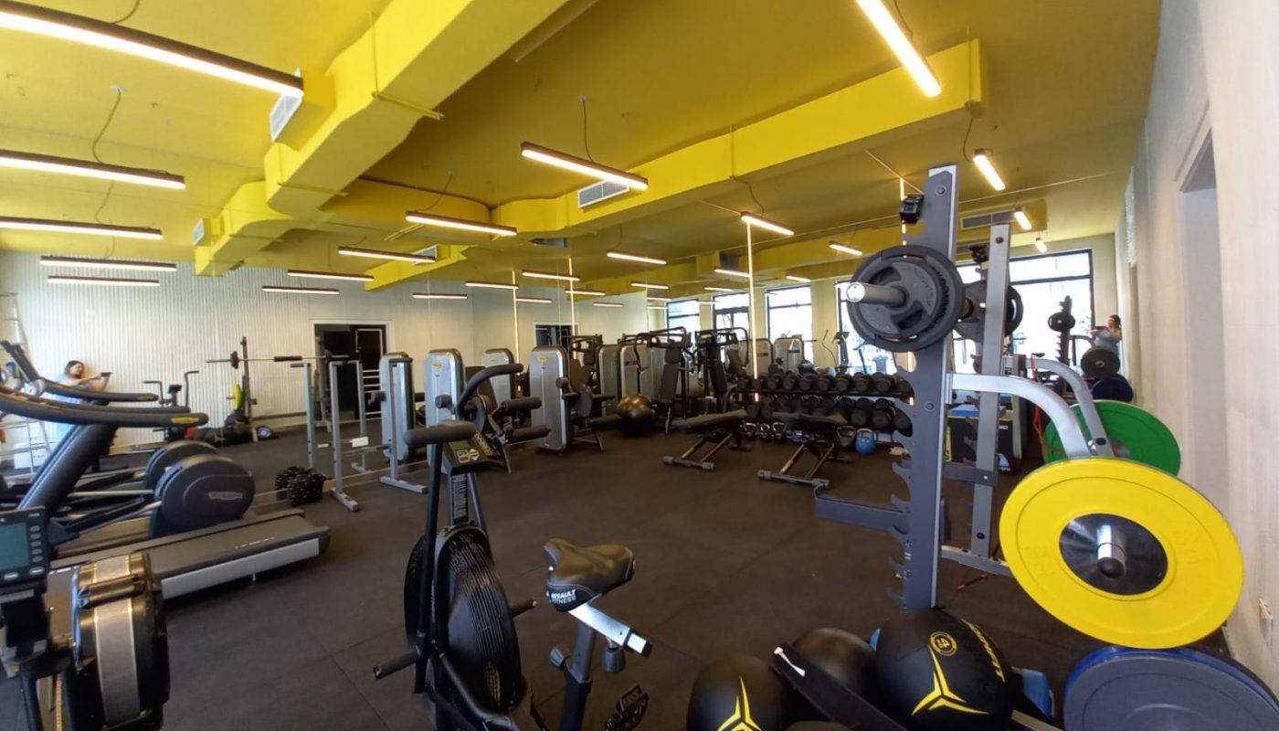 A modern gym with various exercise machines, weights, and equipment under bright yellow ceiling fixtures. No people are present.