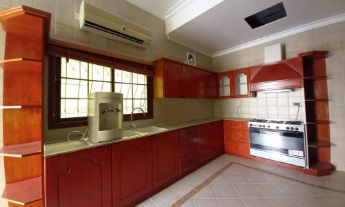 A kitchen with wooden cabinets, built-in stove and oven, a water purifier on the counter, and an air conditioning unit above the window. The floor has a beige and brown tiled pattern.