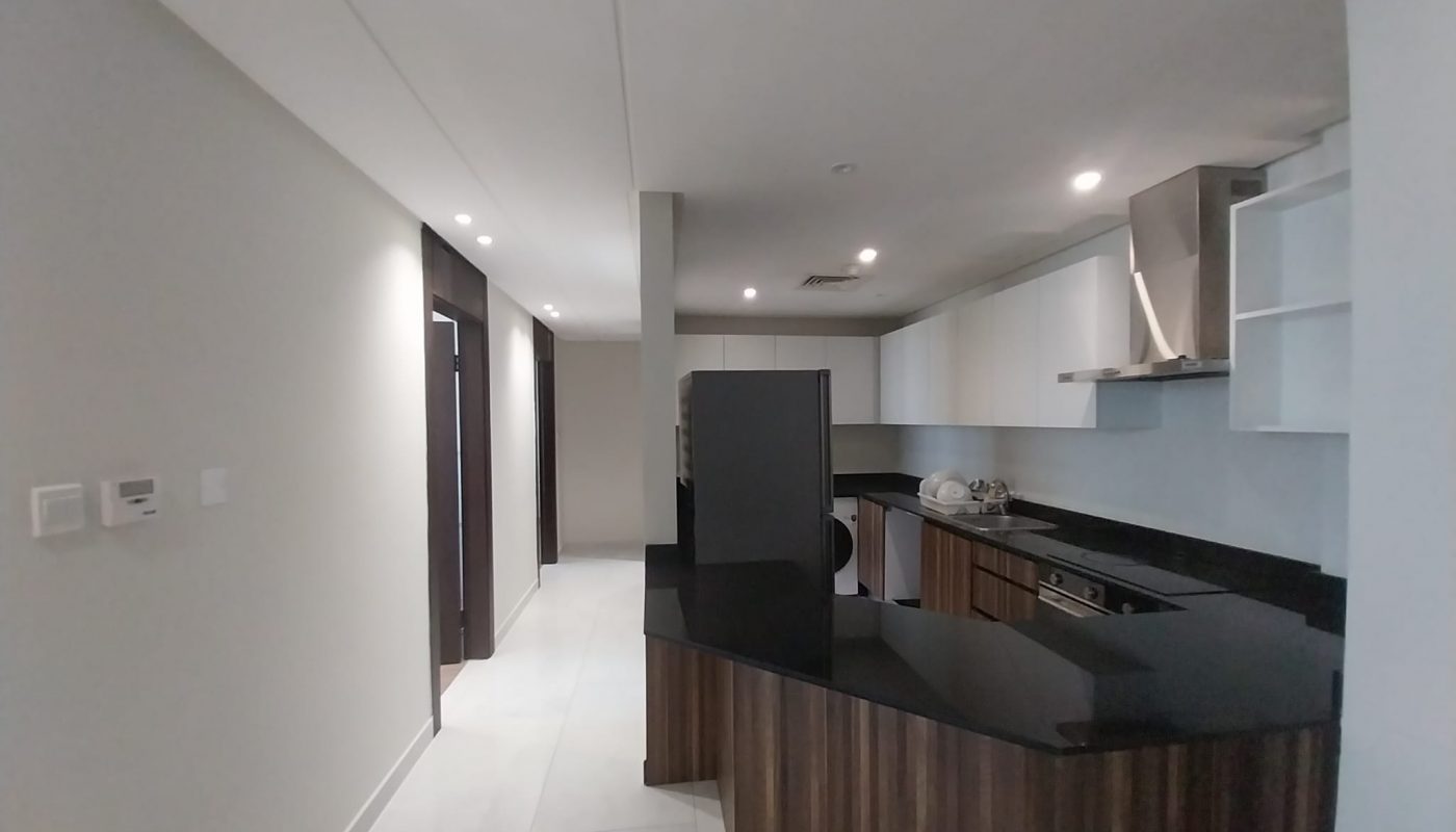 Modern kitchen with black countertops, dark wood cabinets, white upper cabinets, built-in appliances, and white tile flooring, leading to a hallway with closed doors.