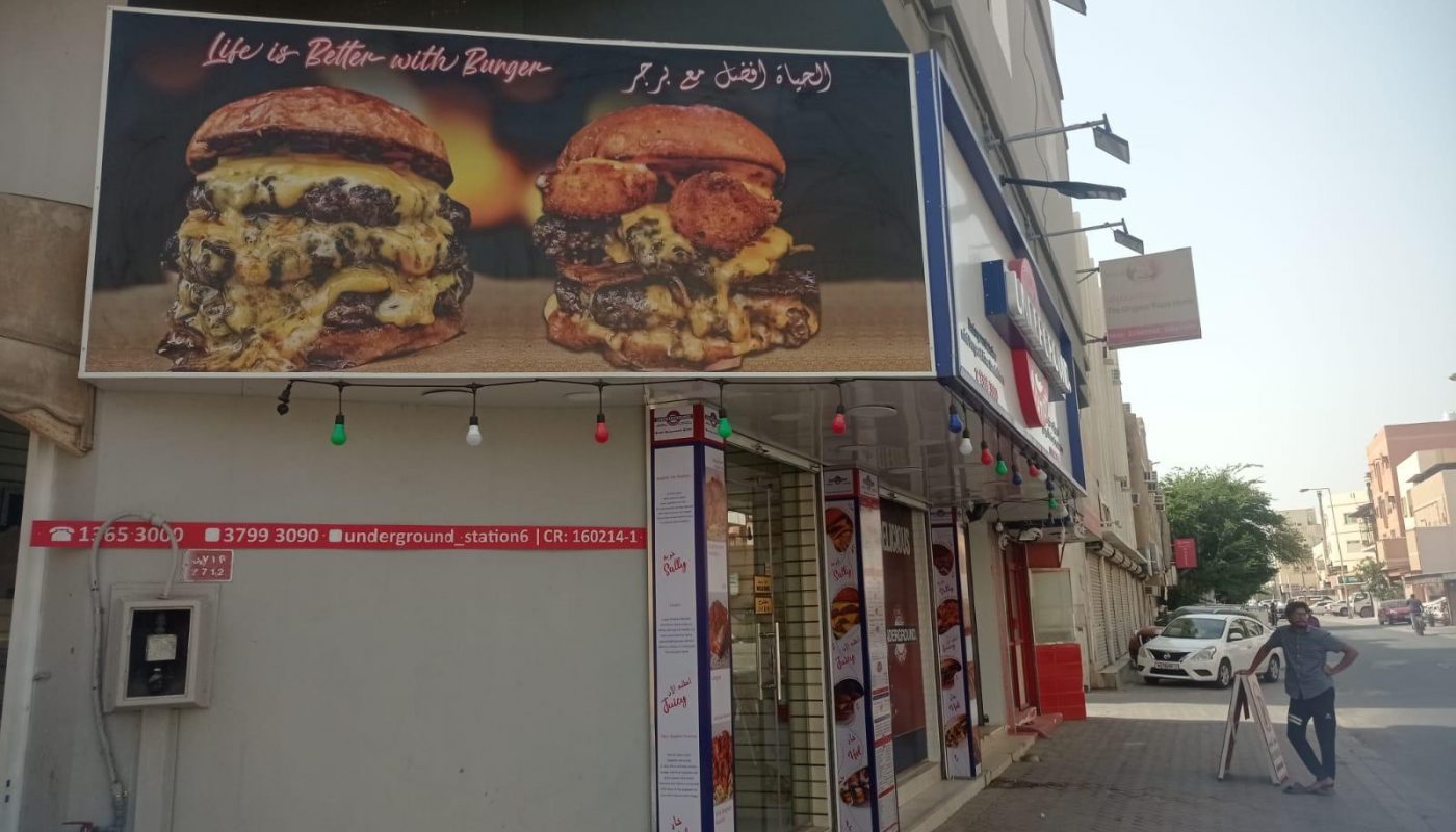 A burger restaurant exterior with a large sign showing two cheeseburgers, menu boards on the windows, and a sidewalk along a city street.