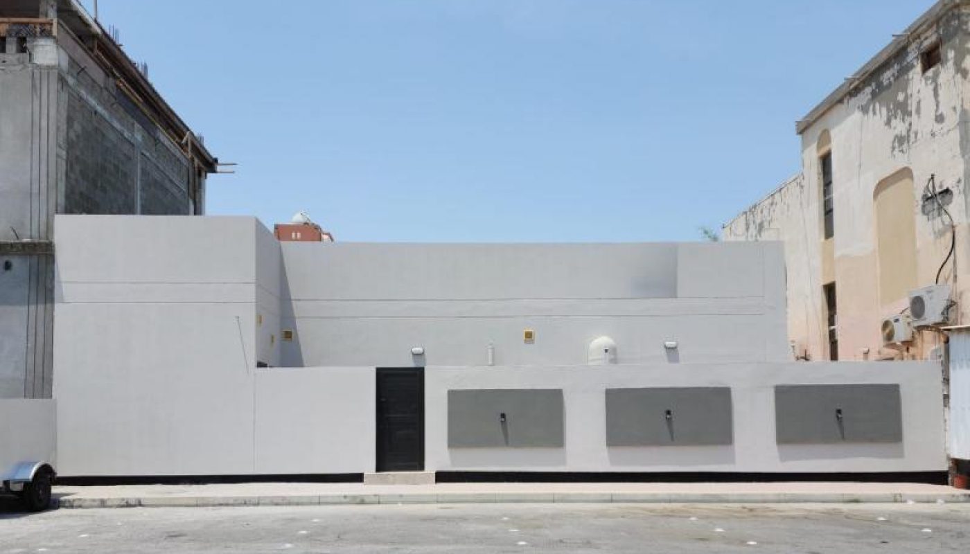 A plain, light gray building with minimal windows and a black door stands between two older, weathered structures under a clear blue sky.