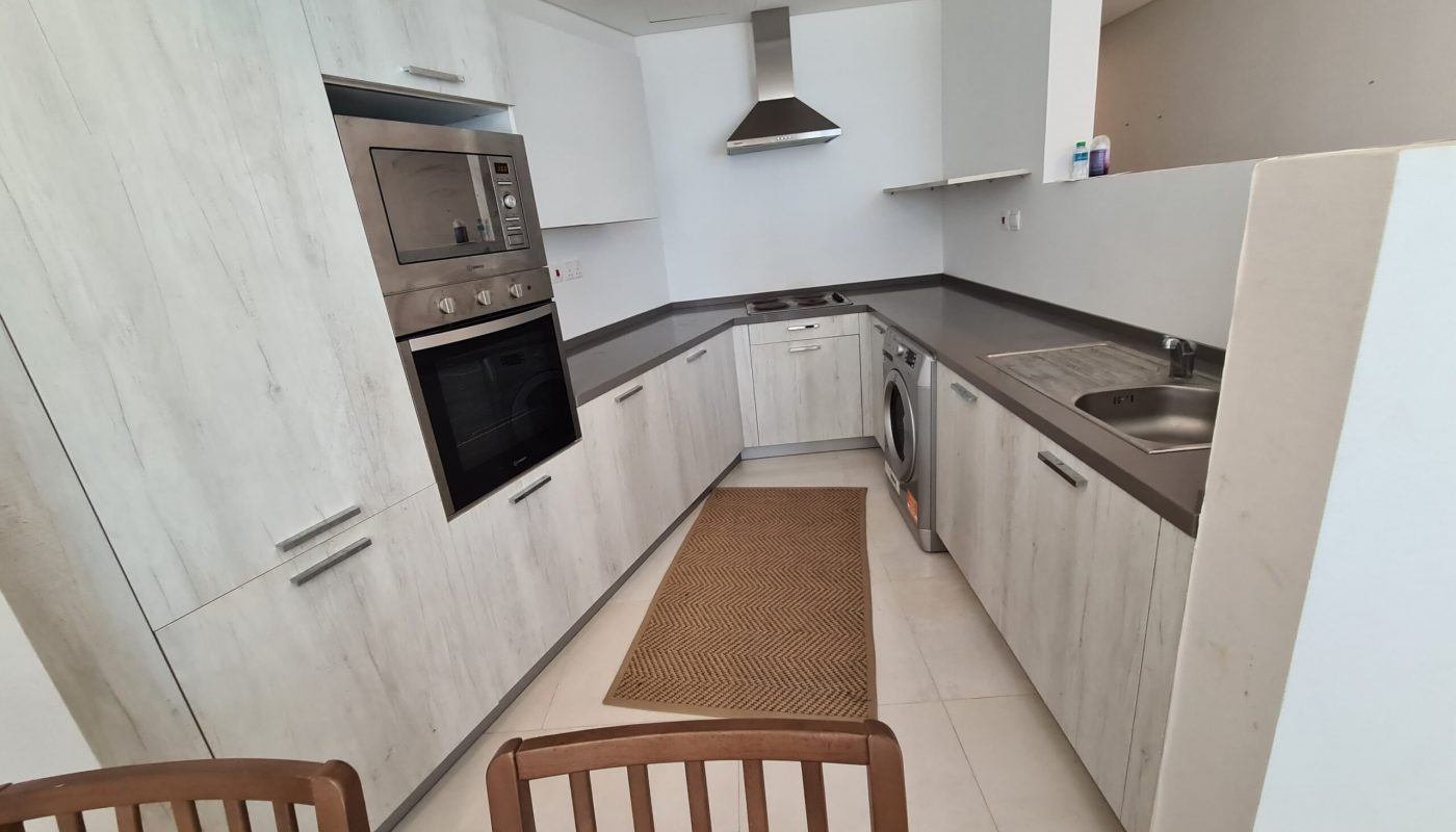 Modern kitchen with light wood cabinets, built-in double oven, stovetop, washing machine, and sink. Brown rug on tiled floor. Two wooden chairs partially visible in the foreground.