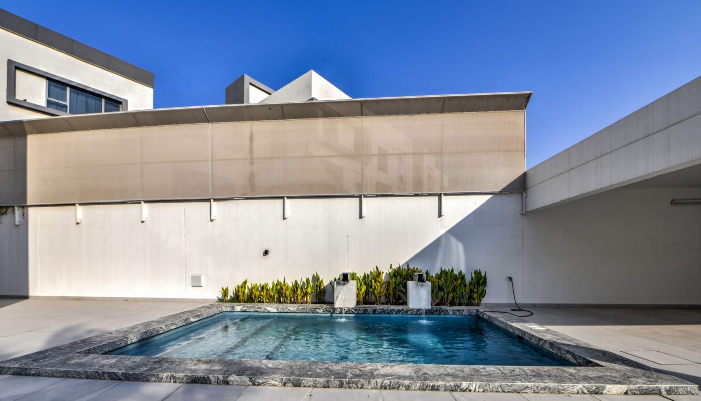 Small rectangular pool with stone edging in a modern, minimalistic courtyard. White walls, potted plants, and clear blue sky in background.
