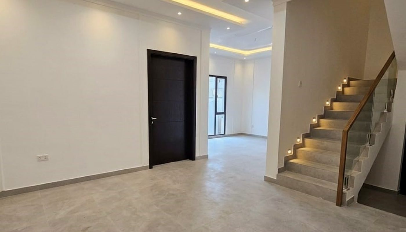 Modern, empty room with tiled flooring, recessed ceiling lights, a dark door, a glass door to the outside, and a staircase with LED step lights and a glass railing.