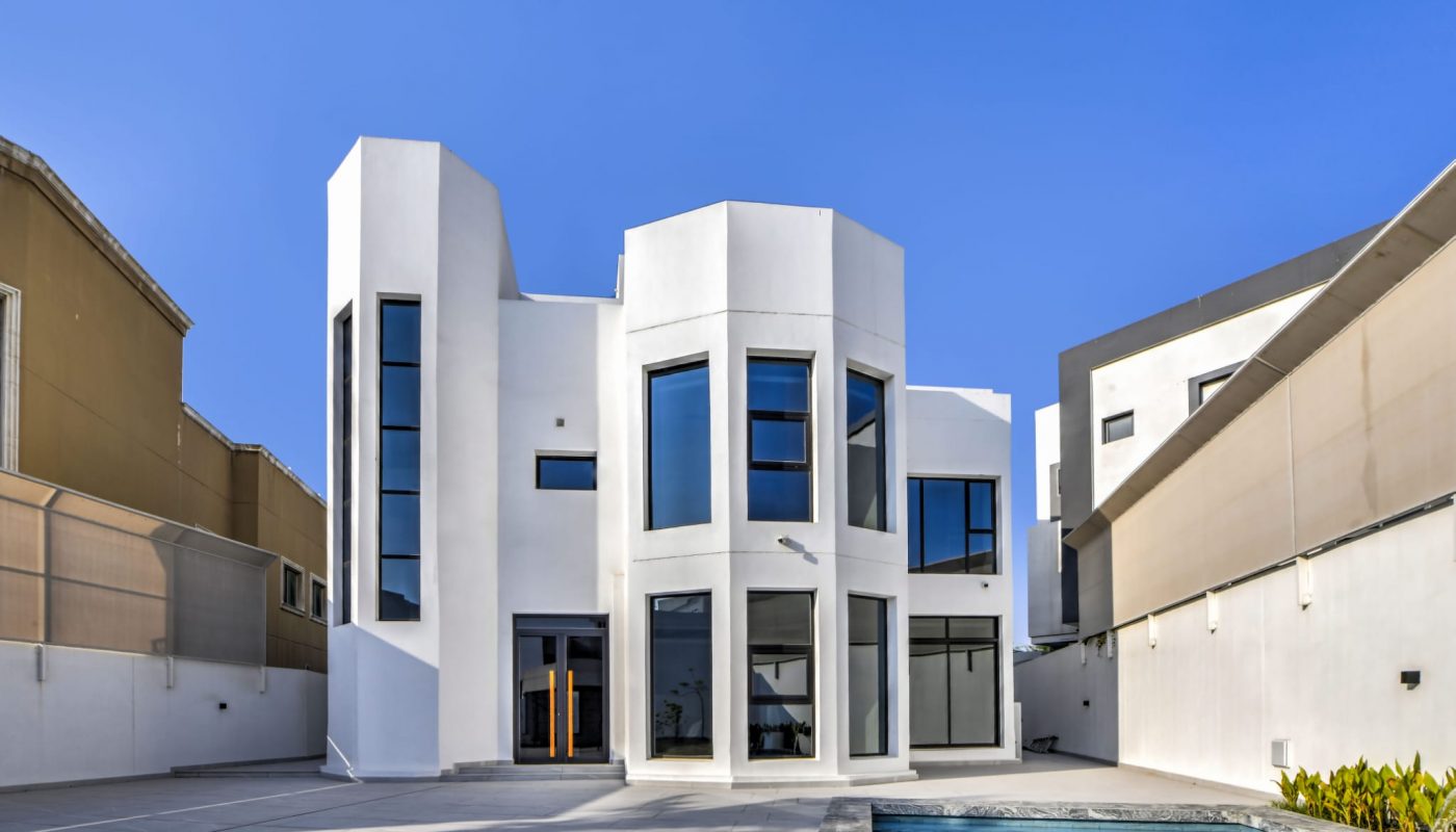 Modern two-story white house with large windows, flat roof, and a small swimming pool in the foreground under a clear blue sky.