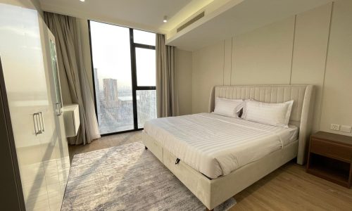 A modern bedroom in this AMWAJ flat for sale boasts a large window, a double bed with white linens, a bedside table, and a wardrobe. The room features wood flooring and has a rug under the bed.