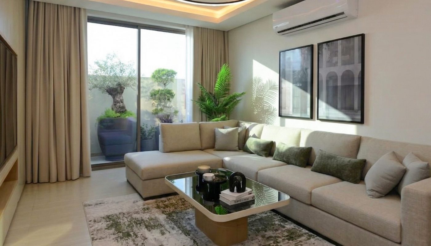 Modern living room with beige sectional sofa, glass coffee table, large rug, wall art, air conditioner, and sliding glass door leading to a balcony with plants.