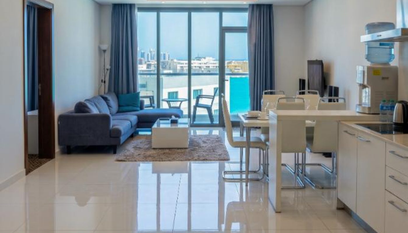 Modern open-plan apartment with white tiled floors, a kitchen and dining area, a gray sofa, and large windows offering a waterfront city view.