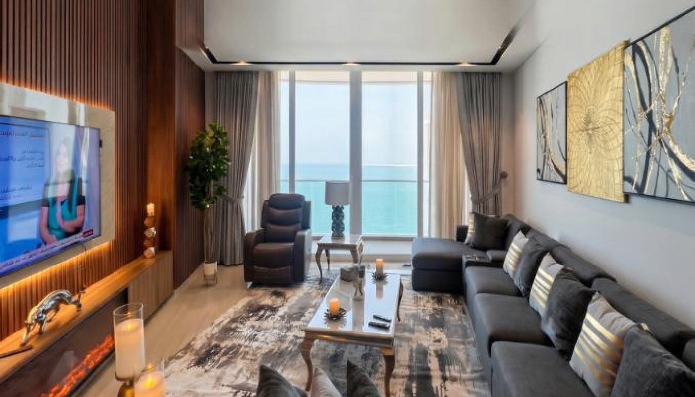 Modern living room with dark furniture, decorative cushions, wall art, TV, and large windows offering an ocean view. Candles and indoor plant add accents to the space.