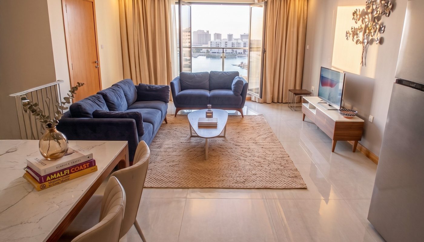 Modern living room with a blue sofa set, coffee table, TV, and large window with city view; dining table and chairs are visible in the foreground.