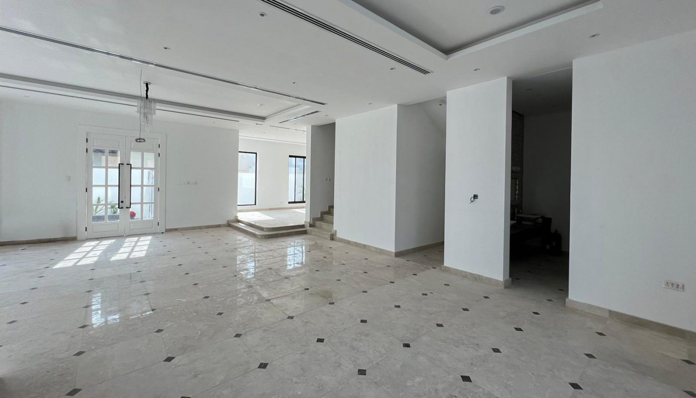 Spacious, unfurnished open-plan room with white walls, tiled floor, large windows, and recessed ceiling lights, featuring a staircase and chandelier.