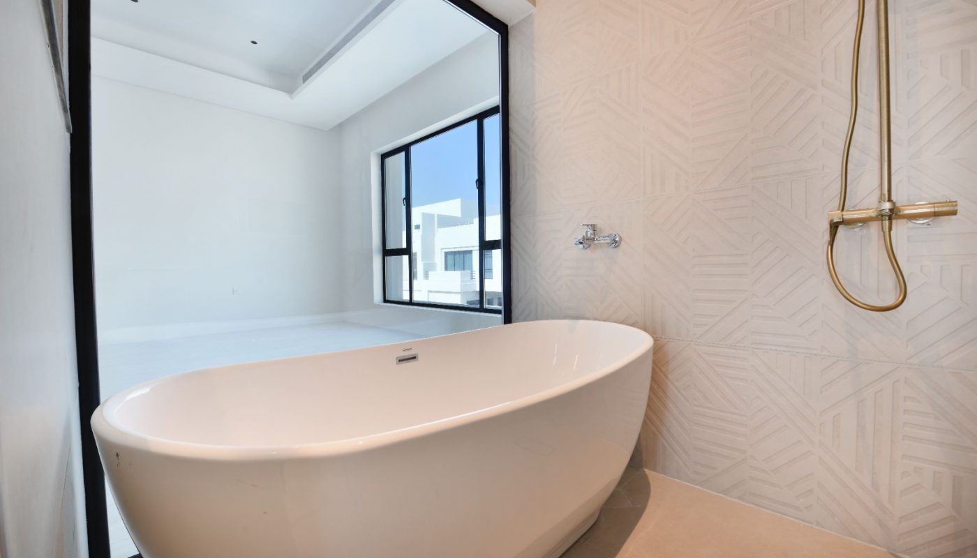 A modern bathroom with a white freestanding bathtub, beige tiled walls, a gold shower fixture, and a large window overlooking a bright room.