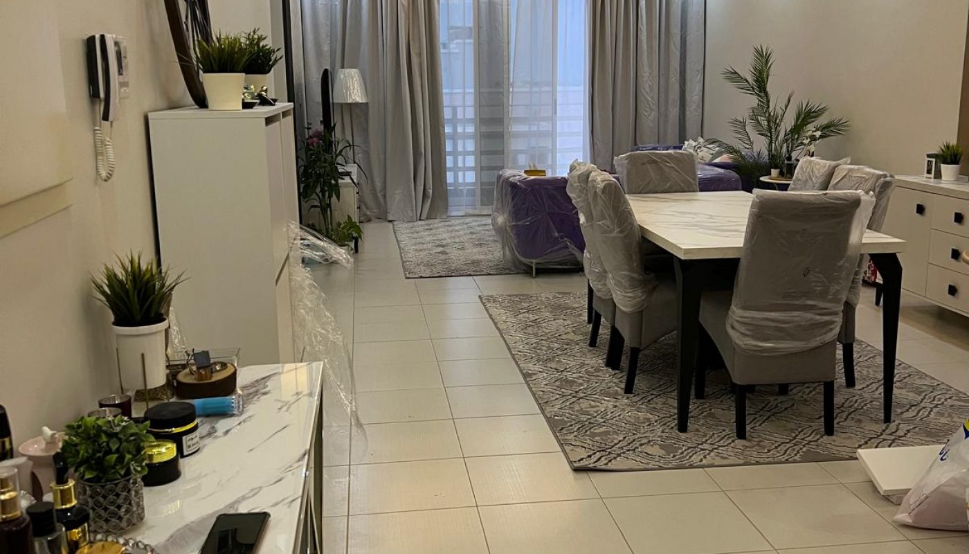 A modern living and dining area with neutral tones, white tiled floor, covered furniture, indoor plants, and a glass door with curtains at the far end.