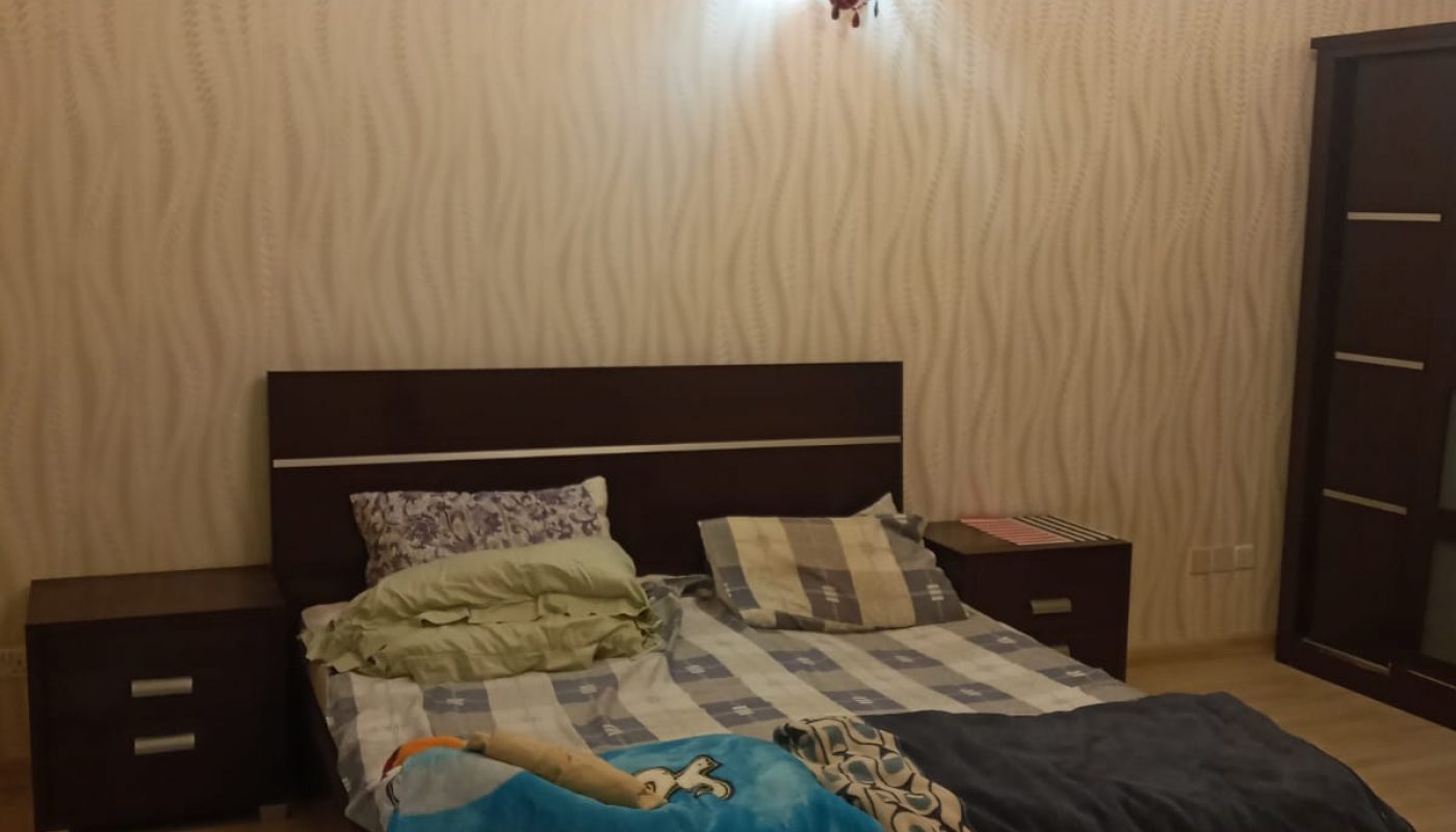 A bedroom with a double bed, unmade bedding, two bedside tables, a wardrobe, and patterned wallpaper, lit by a ceiling light and a red wall lamp.