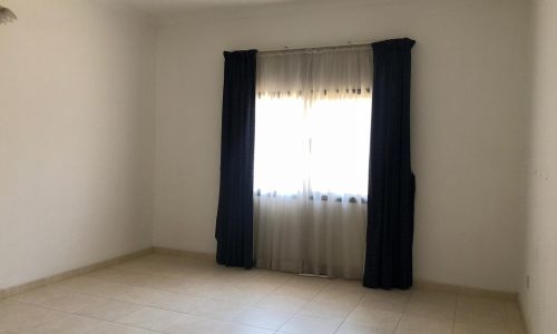 An empty room with beige tiled floors and white walls. A single window with dark curtains and sheer white drapes is centered against the back wall.