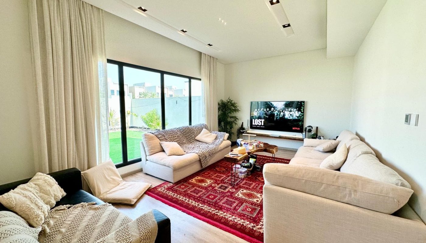 A modern living room with three sofas, a large window, a TV displaying the show 'Lost', patterned rugs, and a coffee table on light wooden flooring.
