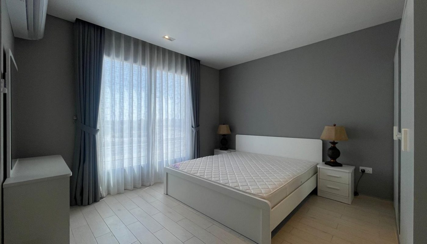 A minimalist bedroom with a double bed, mattress, two bedside tables with lamps, light wood flooring, grey walls, and large window with sheer curtains and dark drapes.