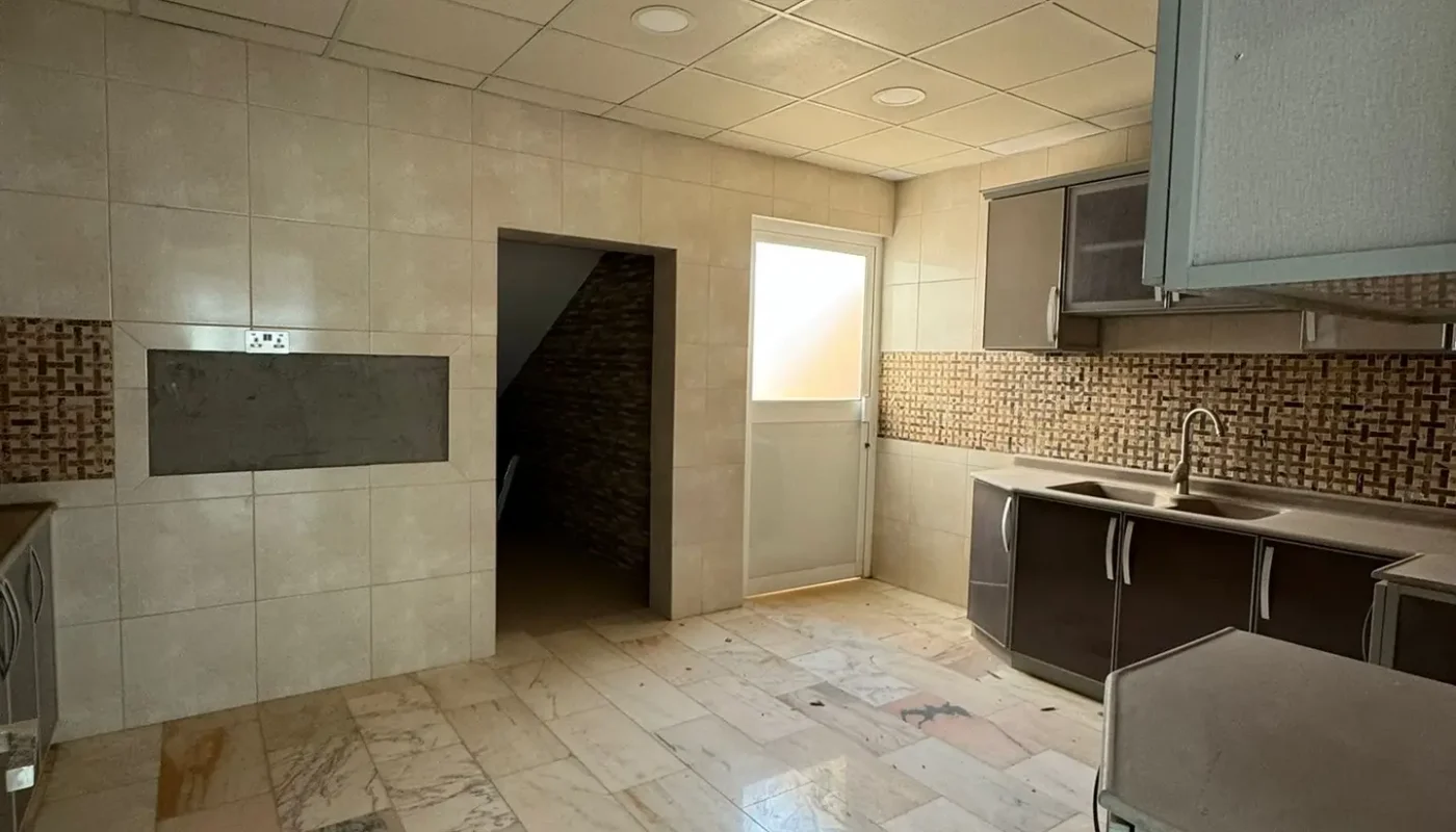 A tiled kitchen with brown cabinets, a sink, and a small open doorway leading to a dark, under-stairs storage area. Natural light enters through a door with frosted glass.